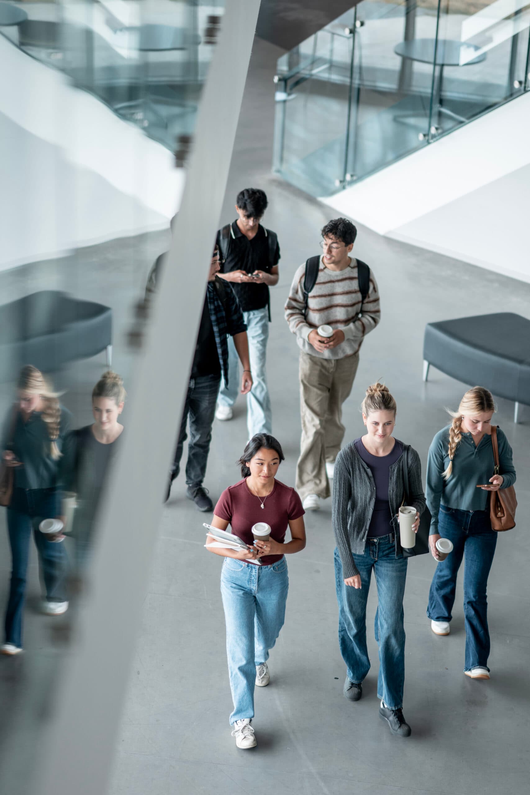 A multi ethnic group of college students walking inside a bright, modern educational building, holding books and coffee, engaging in conversation, and displaying youthful camaraderie and collaboration, symbolizing diversity and education.